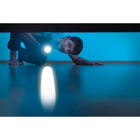 A man crawls under a piece of furniture with a flashlight shining on the floor. Blue wall in the background.