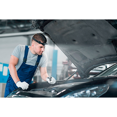 A mechanic with a headlamp looks under the hood of a car, wearing white work gloves.