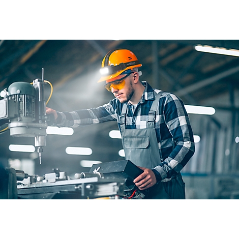 Man in work clothes at a machine. He wears an orange helmet with light, safety glasses. Beam of light.