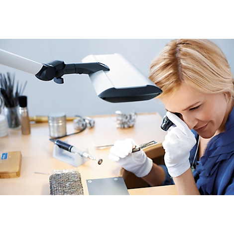 A woman in a blue blouse and gloves inspects a small object with a magnifying glass and tools under a lamp.