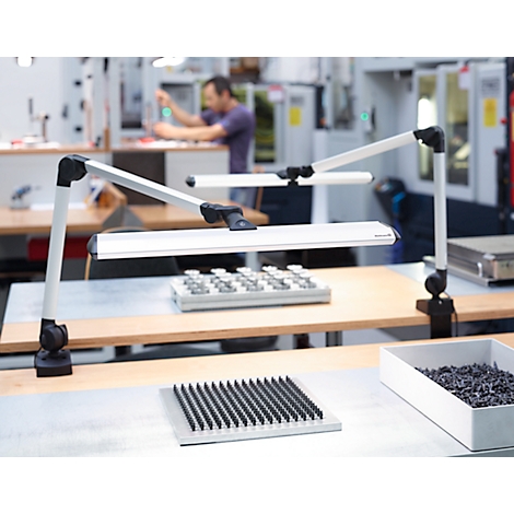 Two desk lamps with swivel arms, in a manufacturing plant. People are working in the background.