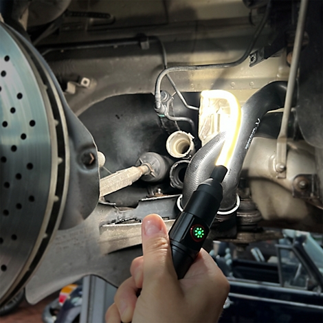 Eine Hand hält eine schwarze LED-Taschenlampe und leuchtet damit in den Motorraum eines Autos. Grüne Leuchte ist an.