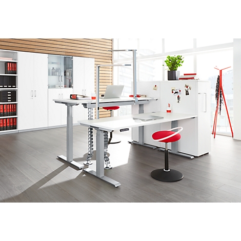 A modern office with height-adjustable desks and a red chair. White cabinets, plants, and a red coat rack complement the image.