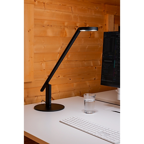 A black desk lamp illuminates a desk with a glass of water, a keyboard, and a computer screen.
