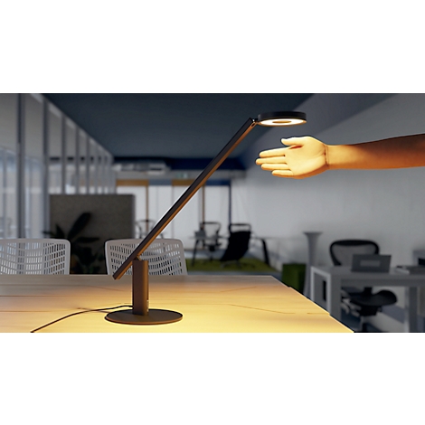A stylish desk lamp with an arm, standing on a table. A hand is approaching the light. In the background an office.