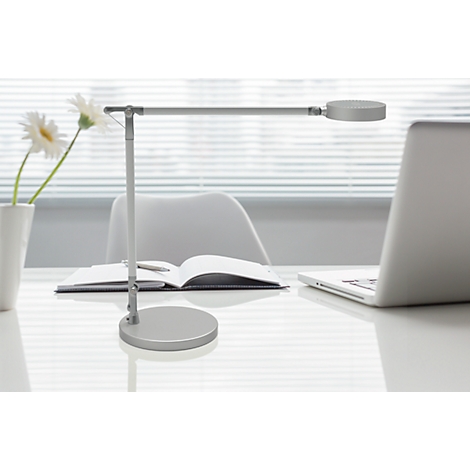Silberne Schreibtischlampe auf Tisch, daneben Laptop, Buch und Vase mit Blumen.