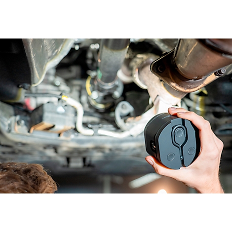 A hand holds a black device under a car. The device appears to be a diagnostic tool.