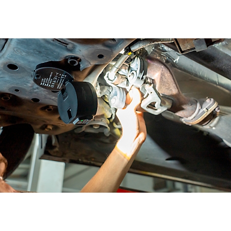 A person working under a car. They are holding a tool near an exhaust pipe. A black device is attached to the car's body.