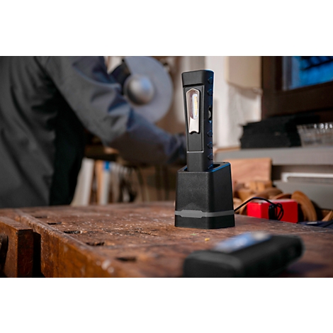 Black LED work light in charging station on wooden surface. In the background, a person is working with tools.