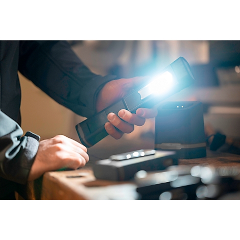 A hand holds a bright flashlight. Tools and a device in the background.