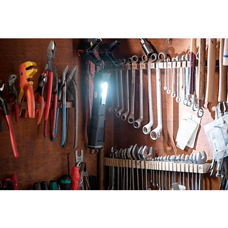 Tools like pliers, wrenches, and hammers hang on a wooden board. A light shines on a cordless screwdriver.