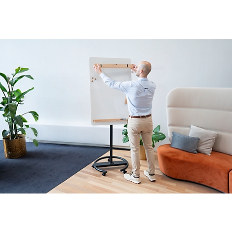 Un homme fixe un tableau blanc. Il se tient devant un fond blanc, d'autres meubles de bureau sont visibles.
