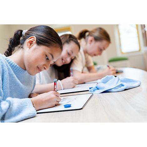 Drei Mädchen schreiben konzentriert mit Stiften auf kleinen weißen Tafeln in einem hellen Klassenzimmer.