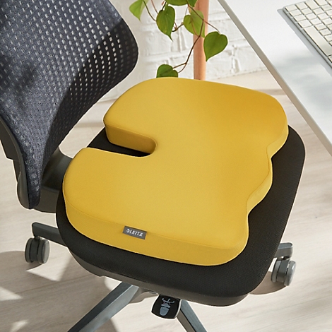 Coussin de siège ergonomique jaune sur une chaise de bureau. Avec découpe et petite étiquette. Fond: chaise, plante, bureau.