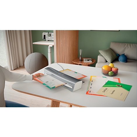 Person laminiert Dokumente auf einem weißen Tisch. Auf dem Tisch liegen weitere Dokumente und eine Obstschale.