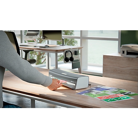 Eine Person legt ein Dokument in ein Laminiergerät auf einem Schreibtisch. Im Hintergrund: Fenster, Monitor, Kopfhörer.