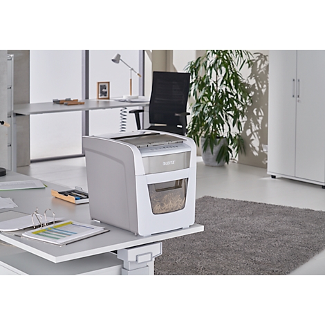 White paper shredder on a desk. In the background an office with plant, desk and cabinet.