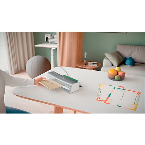 A person places a document into a laminator on a table. There are also laminated cards on the table and a bowl of fruit.