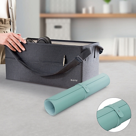 Tapis de bureau bleu clair enroulé avec une lanière en cuir et une boîte de rangement grise. À côté, une main qui touche la boîte.