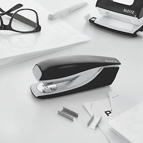 Black stapler with white accents, pens, glasses, hole punch, and paper on a white table. LEITZ logo.