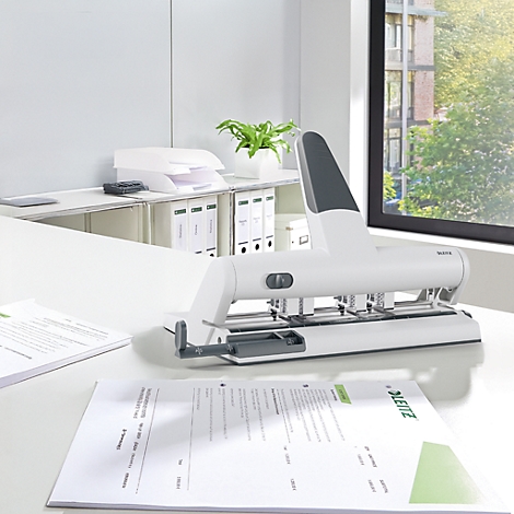 White hole punch on a desk with paper. In the background office furniture and a window with a view of greenery.