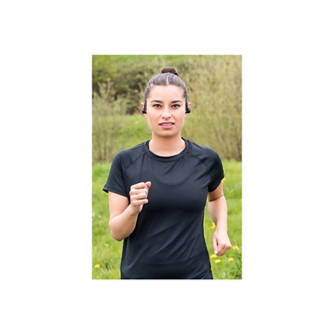 LÃ¤uferin mit KopfhÃ¶rern im schwarzen Shirt. Sie blickt in die Kamera, im Hintergrund Wiese und BÃ¤ume.