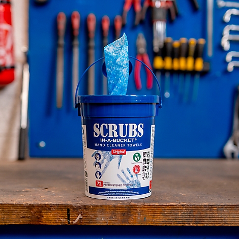 Un seau de lingettes nettoyantes pour les mains sur une surface en bois; des lingettes bleues émergent. Outils en arrière-plan.