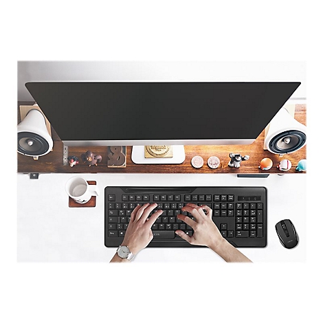 Person tippt auf schwarzer Tastatur. Auf dem Tisch: Monitor, Lautsprecher, Maus, Tasse, Deko.