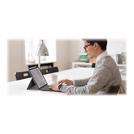 Ein Mann sitzt an einem Tisch und benutzt ein Tablet mit Tastatur. Er trÃ¤gt Brille und grauen Pullover, Kaffee und Lampe.