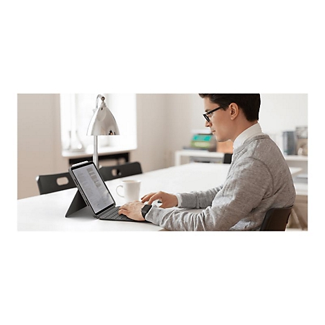 Mann sitzt an Tisch, tippt auf Tablet mit Tastatur. Er trägt Brille, grauen Pullover und weißes Hemd. Auf dem Tisch steht eine Tasse.