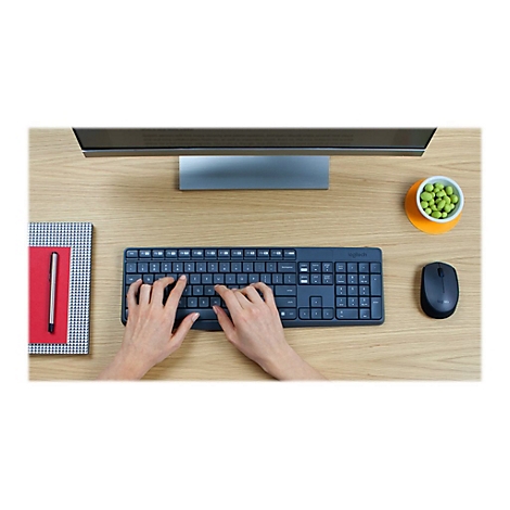 Draufsicht auf Hände beim Tippen auf einer schwarzen Logitech-Tastatur. Auf dem Tisch liegen auch eine Maus, ein Notizbuch und eine kleine Schale mit Erbsen.