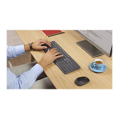 Person tippt auf einer grauen Tastatur. Auf dem Holztisch befinden sich eine Maus, eine Kaffeetasse und ein Monitor.