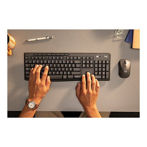 Draufsicht auf eine schwarze Logitech-Tastatur und -Maus, die auf einem grauen Schreibtisch liegen. Die Hände einer Person ruhen auf der Tastatur, eine Uhr am linken Handgelenk.