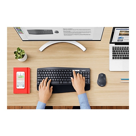 Configuration de bureau avec clavier, souris et smartphone noirs. Des informations s'affichent à l'écran. À côté, un carnet rouge, un moniteur et une petite plante en pot.