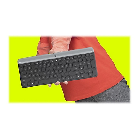 Eine Person hält eine Logitech-Tastatur vor gelbem Hintergrund. Die Tastatur ist grau und schwarz. Im Hintergrund ist ein rotes T-Shirt und eine graue Hose zu sehen.