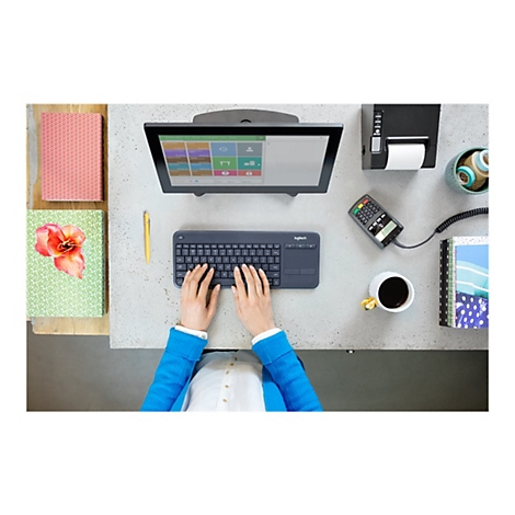 Person bedient Tastatur an einem Schreibtisch, daneben Monitor, Drucker, Kaffeetasse und Notizbuch.