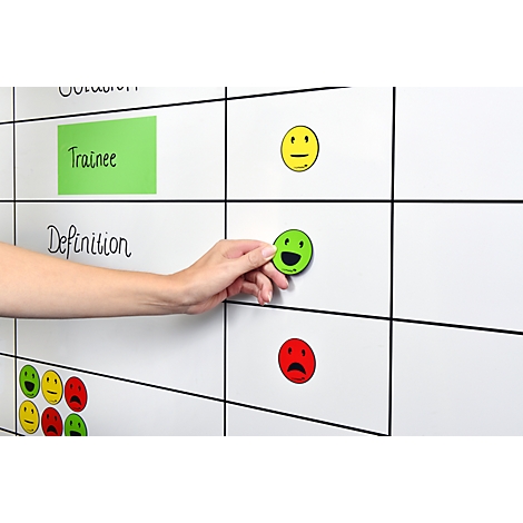 Hand places a green smiley magnet on a white wall. Yellow and red smileys are also visible, along with text like 'Definition' and 'Trainee'.