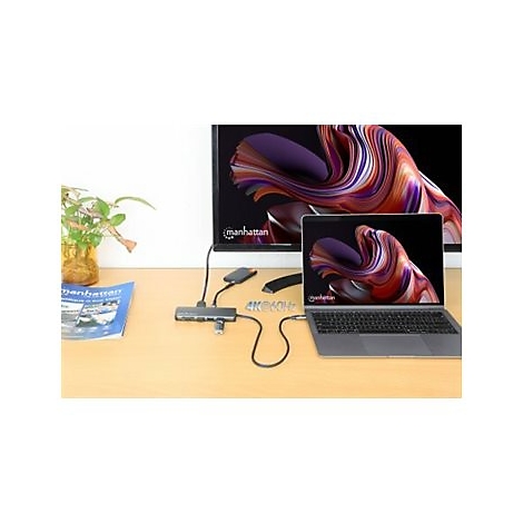 Grauer Laptop mit externem Hub an einem Monitor angeschlossen. Auf dem Tisch liegen Zeitschriften und eine Pflanze.