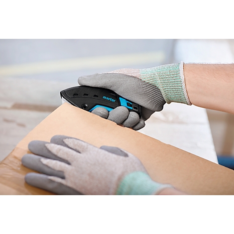 Person schneidet mit einem schwarzen und blauen Cutter Pappe. Hände tragen graue Handschuhe.