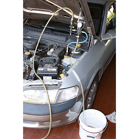 Motor eines silbernen Autos mit geöffneter Motorhaube, verbunden mit Schläuchen und einem Messgerät, Eimer daneben.