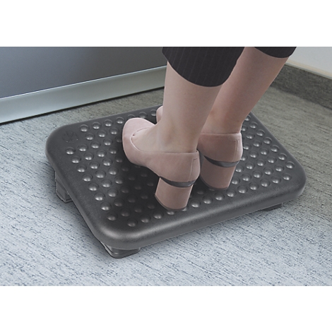 Füße in beigen Schuhen stehen auf einer schwarzen, genoppten Fußstütze unter einem Schreibtisch.