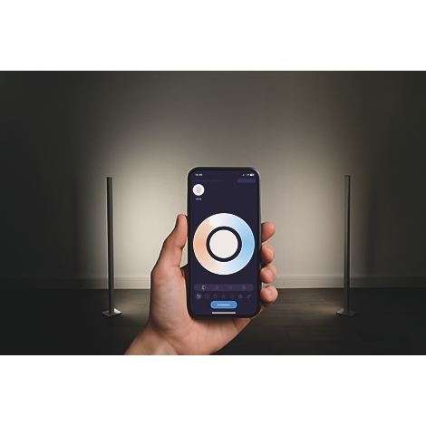 Eine Hand hält Smartphone mit Farbsteuerung. Zwei schlanke Lampen stehen im Raum.