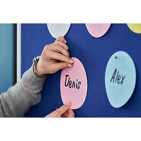 Hand placing a green thumbtack in a pink note with the name "Denis". Blue pinboard in the background.