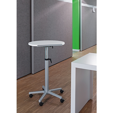 A height-adjustable standing desk with a round, white tabletop stands in an office. The table has a silver frame and castors.