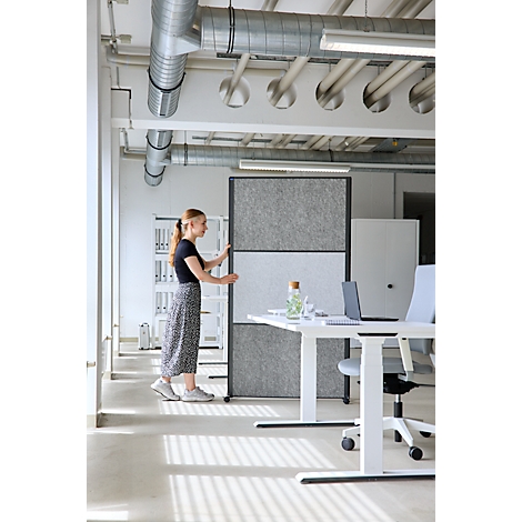Eine junge Frau positioniert einen grauen, dreigeteilten Raumtrenner in einem modernen Büro mit Industrie-Stil.