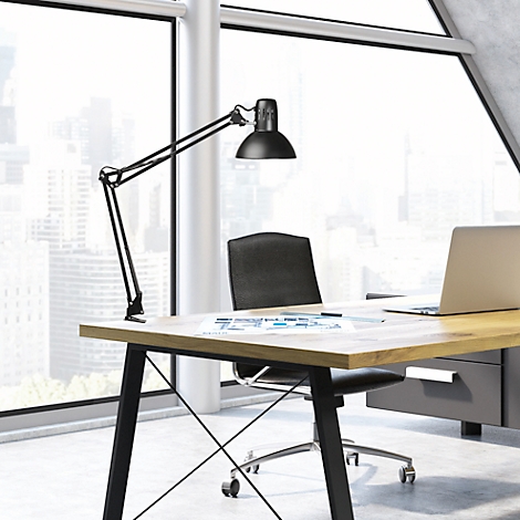 Zwarte bureaulamp boven een houten bureau met laptop en stoel. Ramen op de achtergrond.