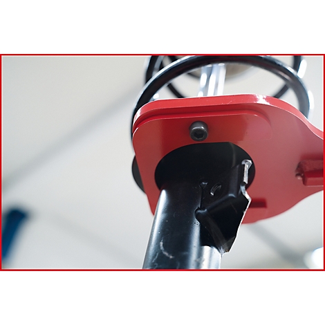 Nahaufnahme eines roten, metallischen Werkzeugs, das an einem Stoßdämpfer befestigt ist. Im Hintergrund ist eine Feder und ein unscharfer heller Raum zu sehen.