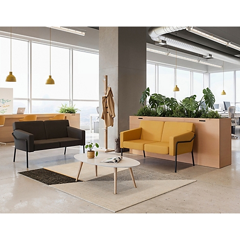 Espace de bureau avec deux canapés, une table basse et des plantes. Canapés jaune et gris devant des fenêtres avec des lampes jaunes.