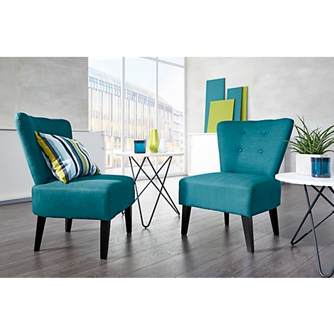 Dos sillones turquesa con patas de madera en un salón luminoso. Un cojín descansa sobre un sillón. En el fondo: ventana, plantas, decoración.