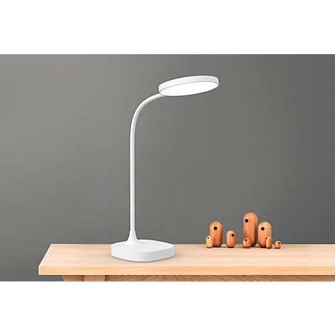 Weiße Schreibtischlampe auf Holztisch, grauer Hintergrund. Holzfiguren daneben.